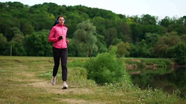 Una chica con una chaqueta rosa y pantalones negros corre cerca del río en auriculares preparándose para la maratón — Vídeos de Stock