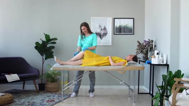 Dos chicas en un salón de belleza maestro en un albornoz y guantes haciendo el procedimiento para eliminar el pelo en las piernas con una mezcla de azúcar — Vídeo de stock