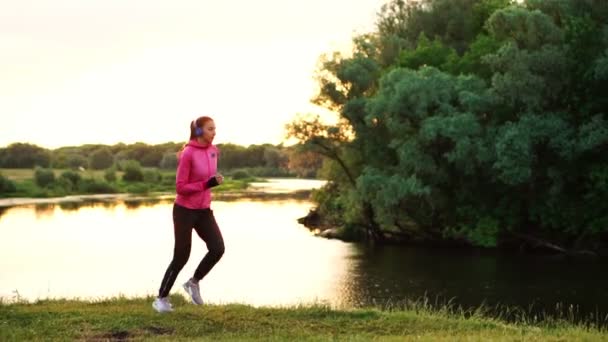 Una chica con una chaqueta rosa y pantalones negros corre cerca del río en auriculares preparándose para la maratón — Vídeos de Stock