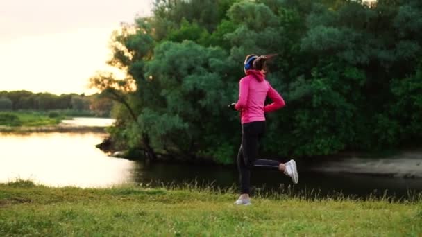 Brunette avec de longs cheveux dans les écouteurs court le long de la rivière dans le parc le matin au lever du soleil en été dans une veste rose et pantalon noir — Video