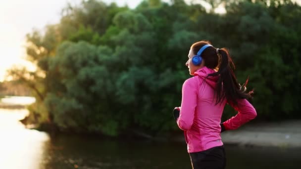 Brunett med långt hår i hörlurar går längs floden i parken på morgonen i soluppgången i sommaren i en rosa jacka och svarta byxor — Stockvideo