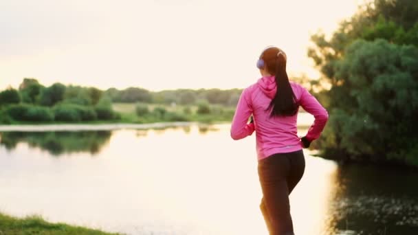 Brunette met lange haren in hoofdtelefoons loopt langs de rivier in het Park in de ochtend bij zonsopgang in de zomer in een roze jas en zwarte broek — Stockvideo