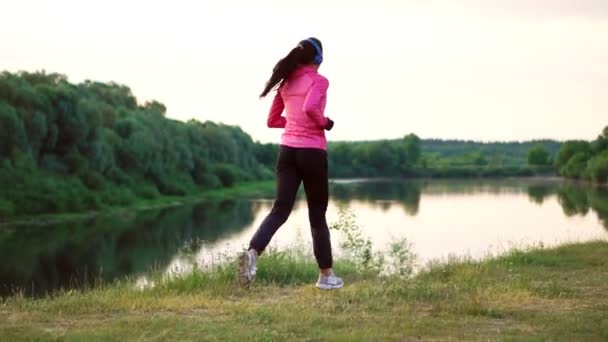Brünette mit langen Haaren und Kopfhörern läuft morgens bei Sonnenaufgang im Sommer in pinkfarbener Jacke und schwarzer Hose im Park am Fluss entlang — Stockvideo