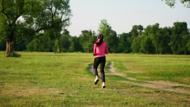 La chica corre al atardecer en el parque a lo largo del estanque y escucha música en los auriculares — Vídeos de Stock