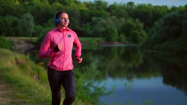 Brunette avec de longs cheveux dans les écouteurs court le long de la rivière dans le parc le matin au lever du soleil en été dans une veste rose et pantalon noir — Video