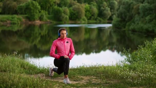 Una chica con una chaqueta rosa se está preparando para un calentamiento y escuchar música en los auriculares a través del teléfono — Vídeos de Stock