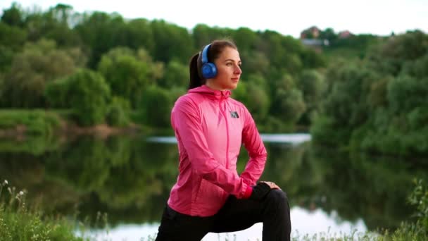 Una chica con una chaqueta rosa se está preparando para un calentamiento y escuchar música en los auriculares a través del teléfono — Vídeo de stock