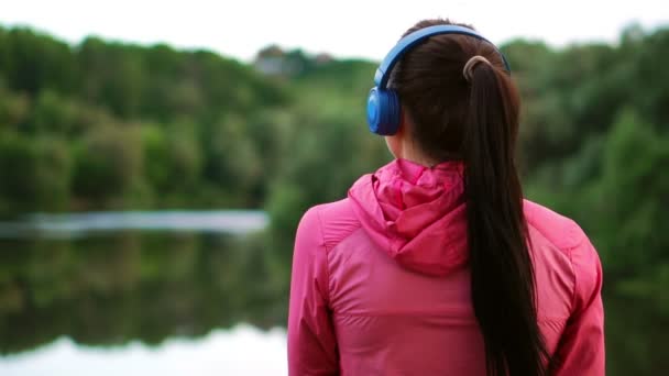 Девушка в розовой куртке и синих наушниках стоит спиной и смотрит на реку рано утром после пробежки — стоковое видео