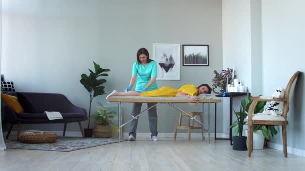 Dos chicas en un salón de belleza maestro en un albornoz y guantes haciendo el procedimiento para eliminar el pelo en las piernas con una mezcla de azúcar — Vídeos de Stock