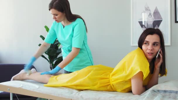 A menina fala ao telefone e sorri durante a depilação das pernas. Shugaring mestre remove o cabelo das pernas — Vídeo de Stock