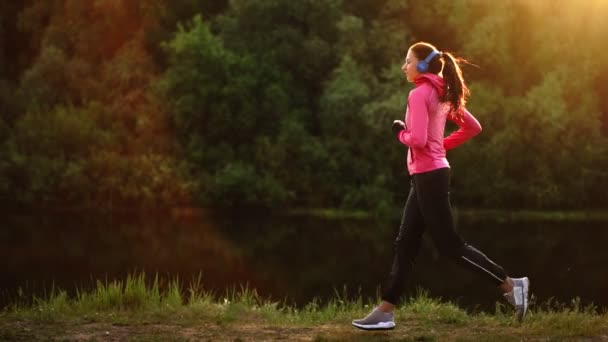 Das Mädchen rennt bei Sonnenuntergang im Park am Teich entlang und hört Musik über Kopfhörer — Stockvideo