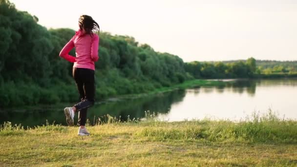 La chica corre al atardecer en el parque a lo largo del estanque y escucha música en los auriculares — Vídeos de Stock