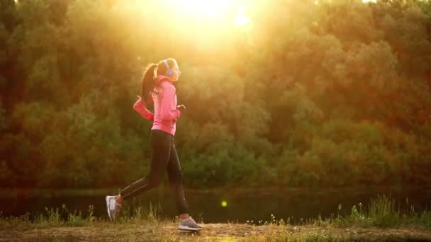 Dziewczyna w różowym kurtkę i czarne spodnie biegnie w pobliżu rzeki w słuchawkach przygotowania do maratonu — Wideo stockowe