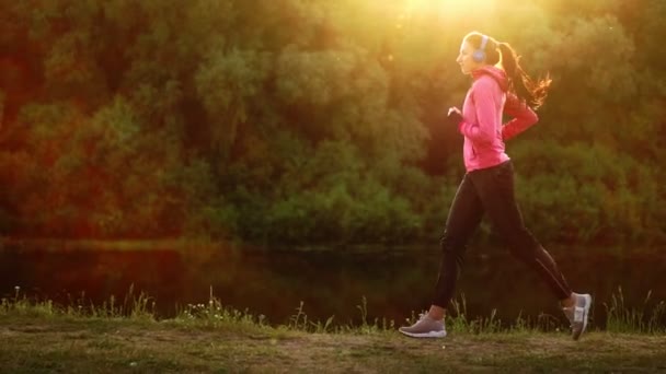 Una chica con una chaqueta rosa y pantalones negros corre cerca del río en auriculares preparándose para la maratón — Vídeos de Stock