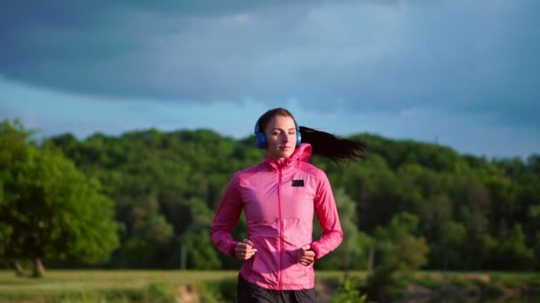 Дівчина в рожевій куртці і чорні штани біжить біля річки в навушниках, готуючись до марафону — стокове відео