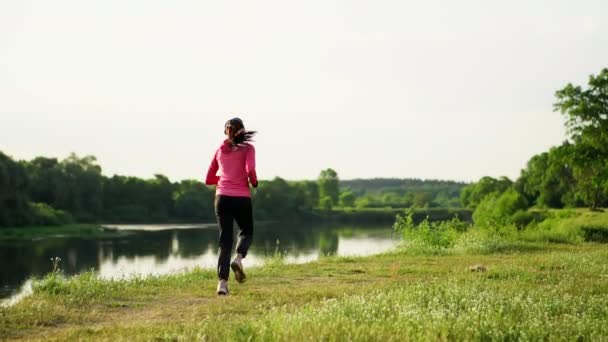 Una chica con una chaqueta rosa y pantalones negros corre cerca del río en auriculares preparándose para la maratón — Vídeo de stock