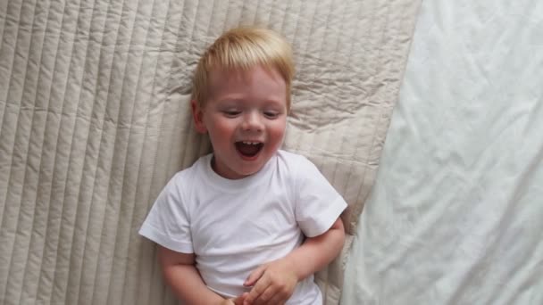 Mom tickles the boy lying on the bed who looks directly into the camera and laughs loudly — Stock Video