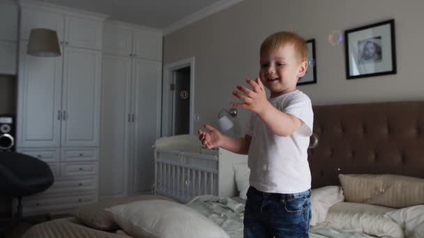 Um menino em uma camiseta branca pula na cama e ri-se ao ver bolhas de sabão no quarto dos pais em câmera lenta — Vídeo de Stock