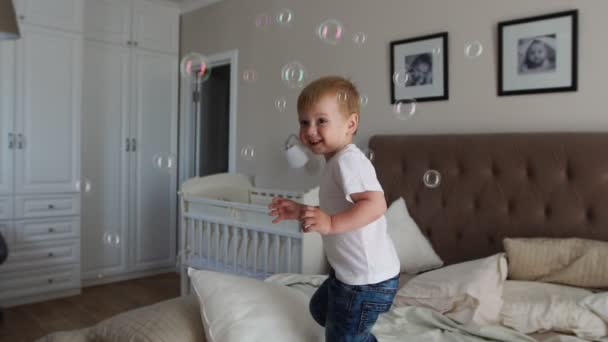 Un garçon dans un T-shirt blanc et un jean bleu attrape des bulles de savon debout sur le lit dans la chambre des parents — Video