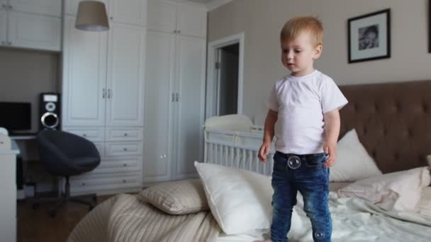 Un garçon dans un T-shirt blanc et un jean bleu attrape des bulles de savon debout sur le lit dans la chambre des parents — Video