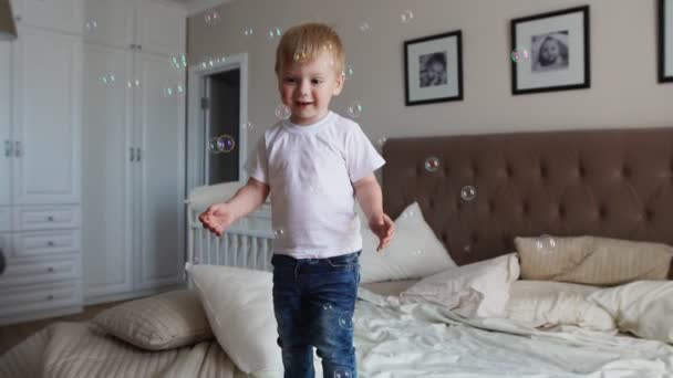 Ein Junge in weißem T-Shirt und blauer Jeans fängt Seifenblasen auf, die auf dem Bett im Elternschlafzimmer stehen — Stockvideo