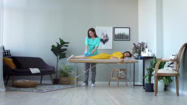 Dos chicas en un salón de belleza maestro en un albornoz y guantes haciendo el procedimiento para eliminar el pelo en las piernas con una mezcla de azúcar — Vídeo de stock