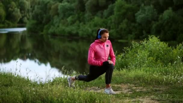 Une fille dans une veste rose se prépare pour une course échauffer et écouter de la musique dans les écouteurs à travers le téléphone — Video