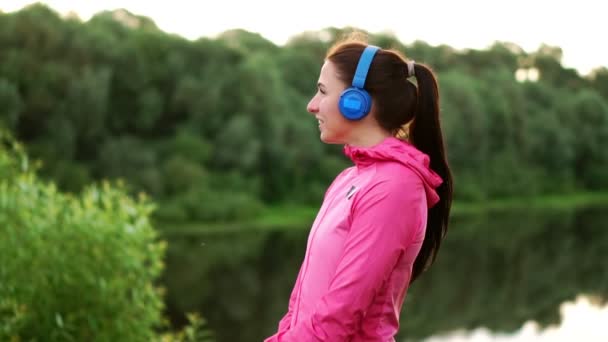 En flicka i en rosa jacka och blå hörlurar står med ryggen och tittar på floden tidigt på morgonen efter en körning — Stockvideo