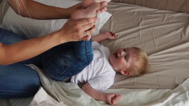 Mama kitzelt den auf dem Bett liegenden Jungen, der direkt in die Kamera schaut und laut lacht — Stockvideo