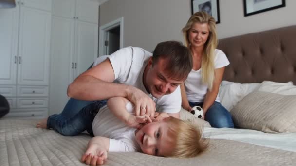 A mãe e o pai fazem cócegas ao filho. pessoas, família, diversão e conceito da manhã - criança feliz com os pais fazendo cócegas na cama em casa. Família feliz passar o tempo juntos na hora de dormir brincando e abraçando — Vídeo de Stock