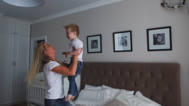 Mom and son are playing on the bed in a bright bedroom, a boy of two years laughs and smiles — Stock Video