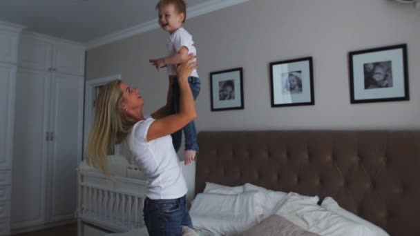 Mãe e filho estão brincando na cama em um quarto brilhante, um menino de dois anos ri e sorri — Vídeo de Stock