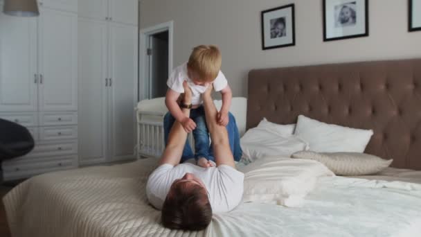 O pai mantém a dignidade acima de si mesmo deitado na cama. Um menino de t-shirt branca ri e sorri de brincar com seu pai — Vídeo de Stock