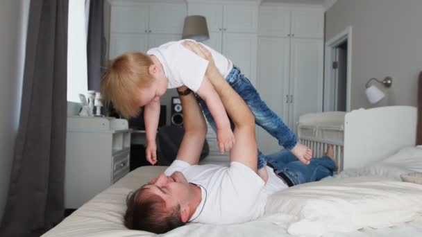Папа держит достоинство выше себя, лежа на кровати. Мальчик в белой футболке смеется и улыбается, когда играет с отцом. — стоковое видео