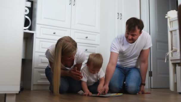 Семья из трех человек в белых футболках и синих джинсах, сидящих на полу своей спальни и играющих с мальчиком в интеллектуальные игры. Медленная съемка счастливая семья — стоковое видео