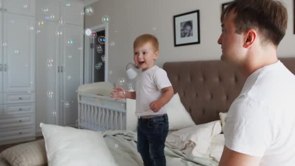 Padre cariñoso juega hijo mirando burbujas de jabón, el niño es feliz saltando en la cama — Vídeos de Stock