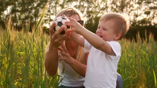 El concepto de una familia feliz. Primer plano de un niño y su madre en un campo con espigas de trigo sonriendo y jugando con una pelota de fútbol — Vídeo de stock