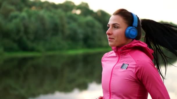 A girl in a pink jacket and black pants runs near the river in headphones preparing for the marathon — Stock Video