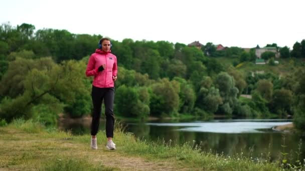Девушка в розовой куртке и черных штанах бежит возле реки в наушниках, готовясь к марафону — стоковое видео
