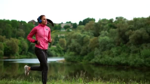 Una chica con una chaqueta rosa y pantalones negros corre cerca del río en auriculares preparándose para la maratón — Vídeo de stock