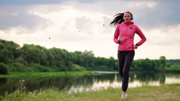 Una chica con una chaqueta rosa y pantalones negros corre cerca del río en auriculares preparándose para la maratón — Vídeo de stock