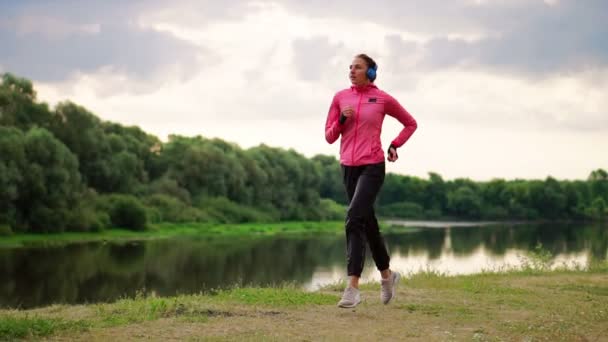 Девушка в розовой куртке и черных штанах бежит возле реки в наушниках, готовясь к марафону — стоковое видео