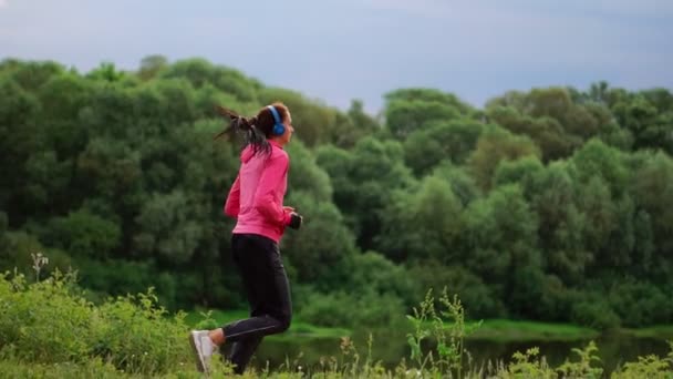 Una chica con una chaqueta rosa y pantalones negros corre cerca del río en auriculares preparándose para la maratón — Vídeos de Stock