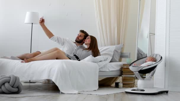 Молодые родители сидят на кровати в белой спальне и делают селфи со спящим ребенком — стоковое видео