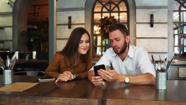 Entusiasmado casal lendo e-mail com boas notícias em um smartwatch em um café com uma janela no fundo com uma luz do pôr do sol ao ar livre — Vídeo de Stock