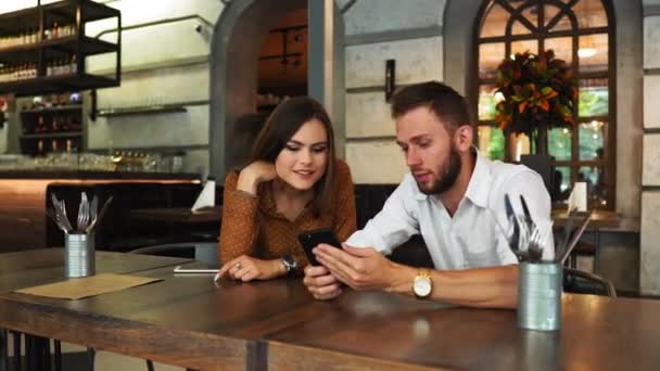 Homme élégant dans les lunettes et belle fille passer du temps ensemble au café, regarder un film sur smartphone en headhone, boire du café avec dessert. Temps avec plaisir . — Video