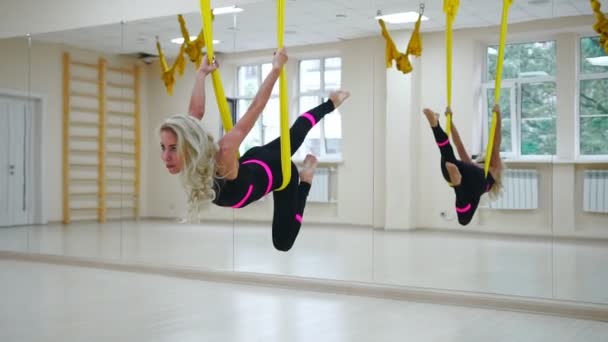 A menina está envolvida em Aero ioga em um estúdio branco voando sobre o chão nas telas em câmera lenta. relaxamento e relaxamento através do alongamento — Vídeo de Stock