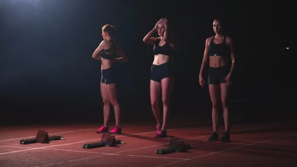 Tres chicas deportivas vestidas de negro del atleta por la noche en la cinta de correr comenzarán la carrera a la distancia del sprint de la posición sentada . — Vídeos de Stock