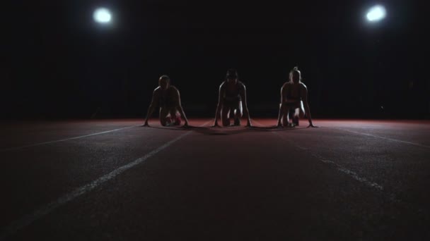 Três meninas em roupas pretas estão nas almofadas iniciais para iniciar a corrida na competição à luz das luzes e correr para o final — Vídeo de Stock