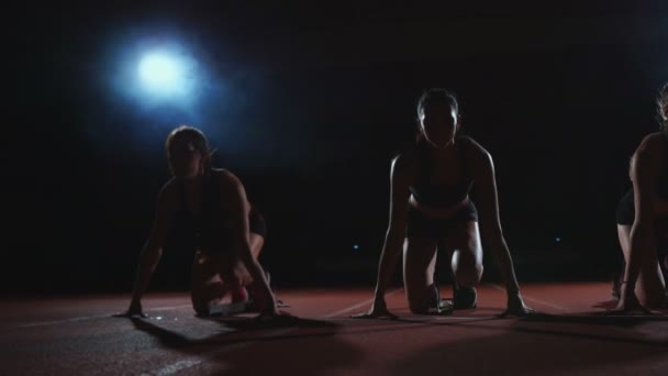 Tres chicas vestidas de negro están en las almohadillas de salida para comenzar la carrera en la competencia a la luz de las luces y correr hacia la meta — Vídeos de Stock